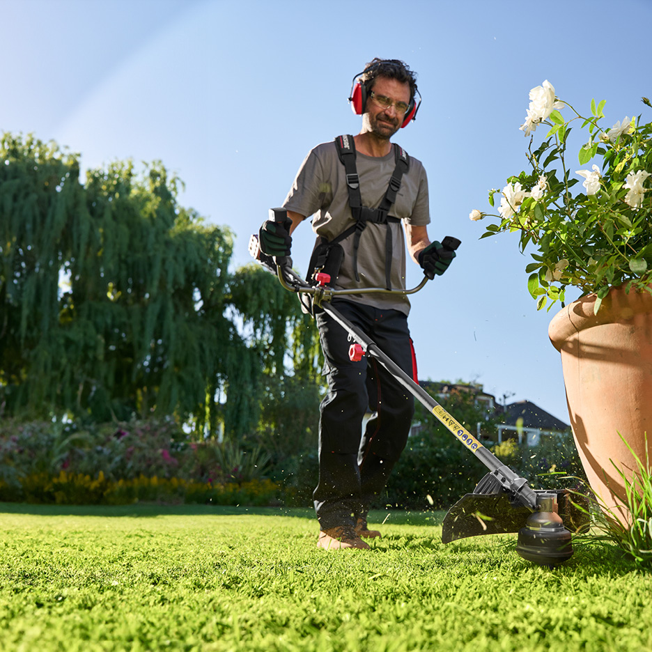 Man using a grass trimmer in a garden, wearing safety headphones and harness.