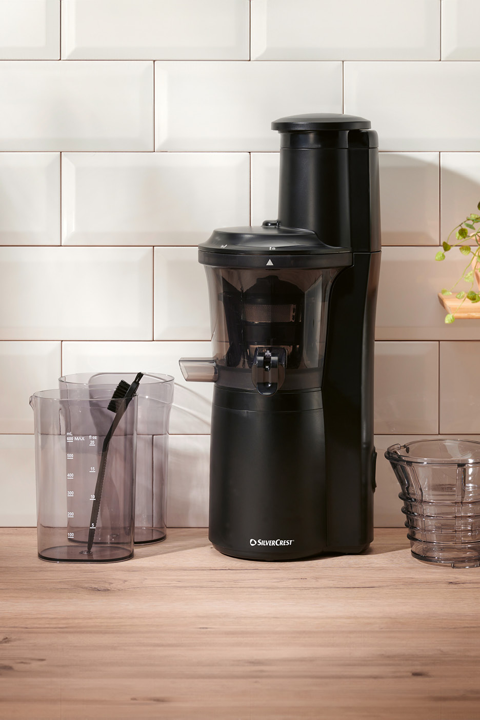 Black slow juicer with two measuring cups and a pulp container on a wooden counter.