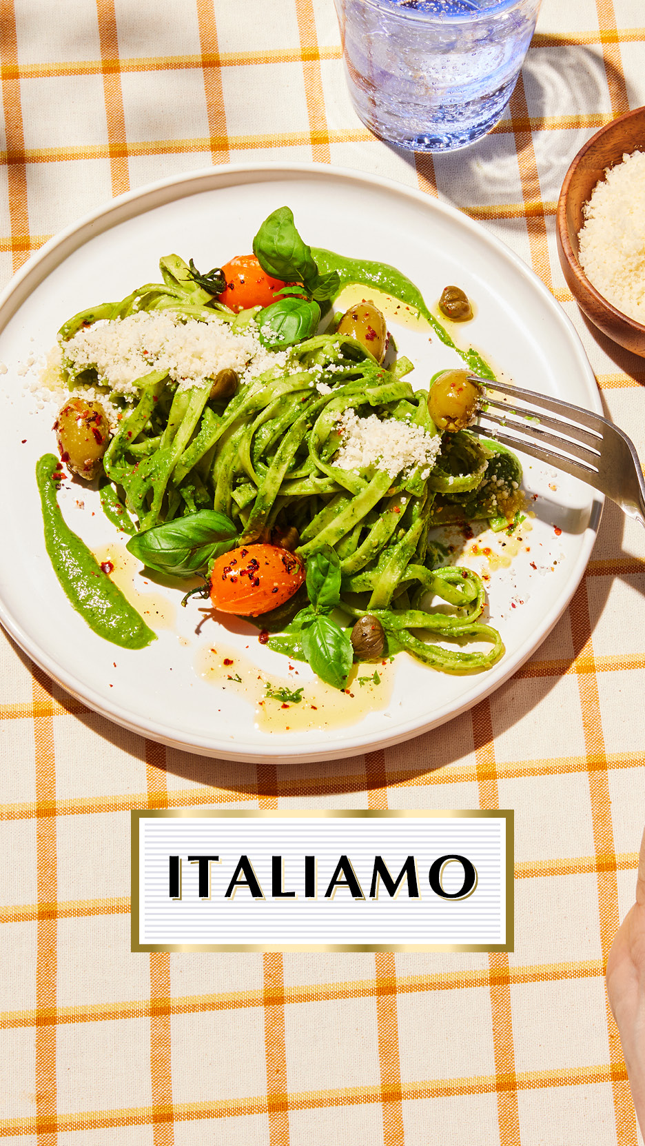 Green pesto pasta with tomatoes, olives, and parmesan on a checkered tablecloth, with the text 'ITALIAMO'.
