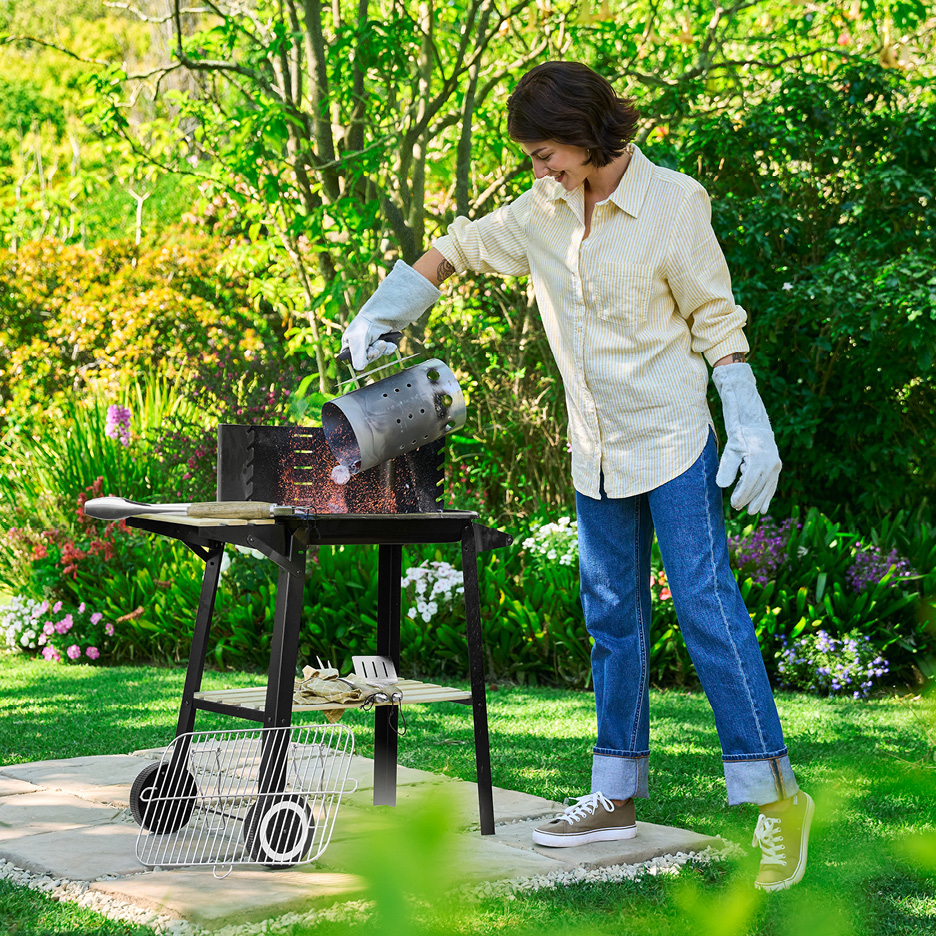 A woman in a garden using a charcoal starter to pour hot coals into a barbecue grill.
