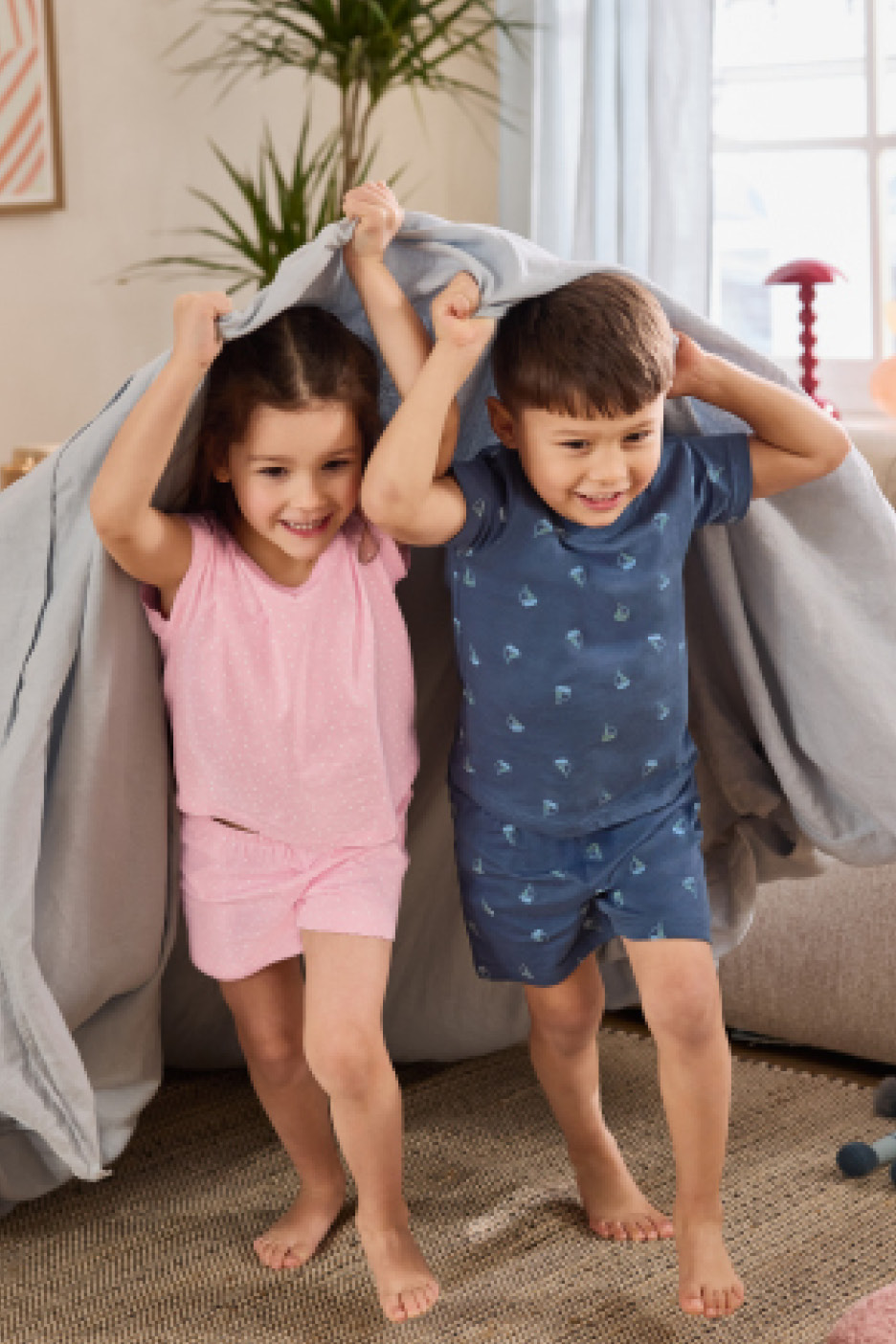 Two children in pajamas playing under a blanket, smiling.