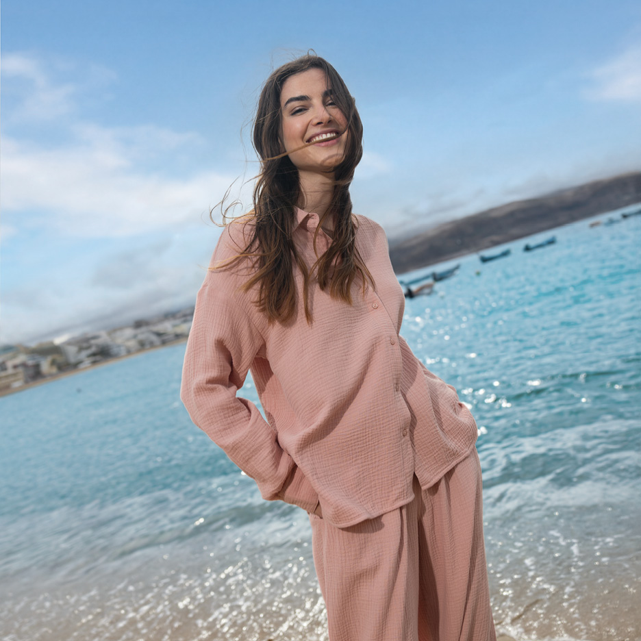 Woman in a pink muslin shirt and trousers on a beach.