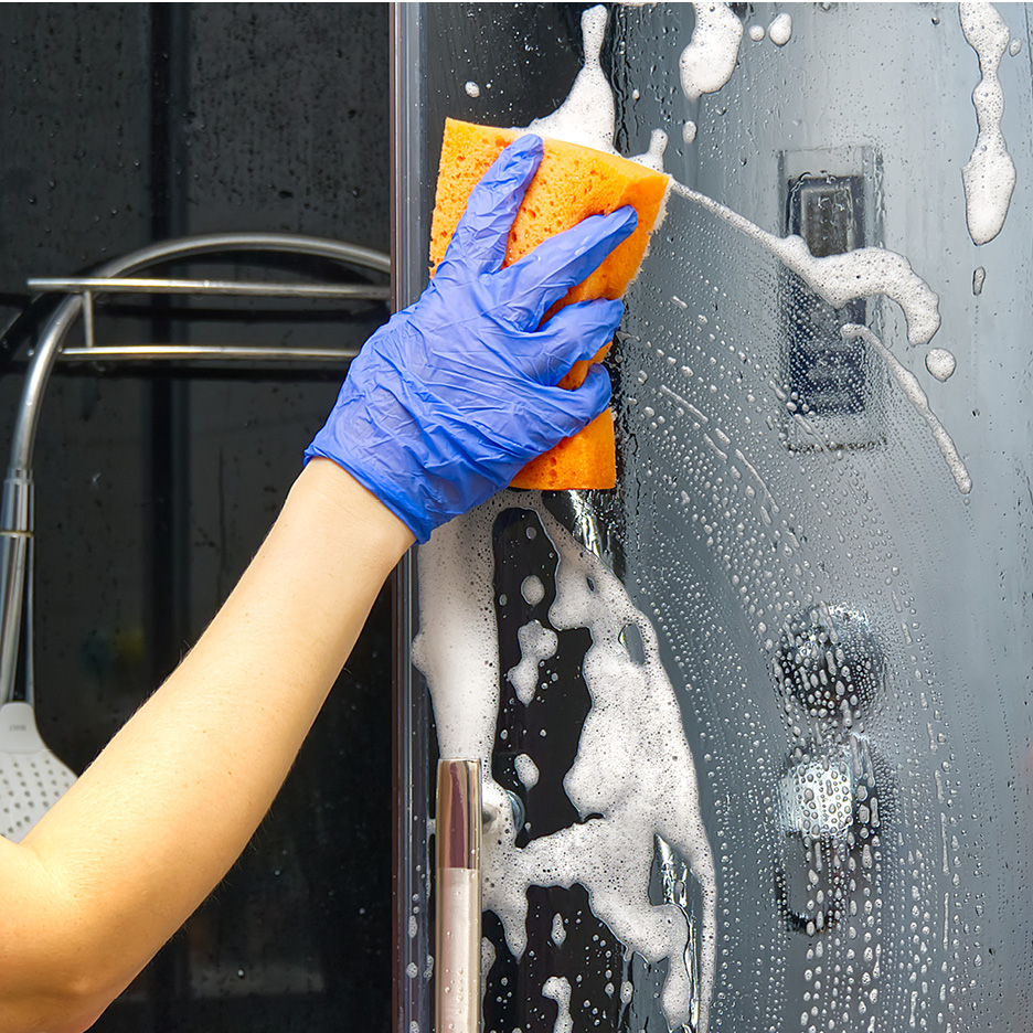 A hand in a blue glove cleaning a shower door with an orange sponge and soap suds.