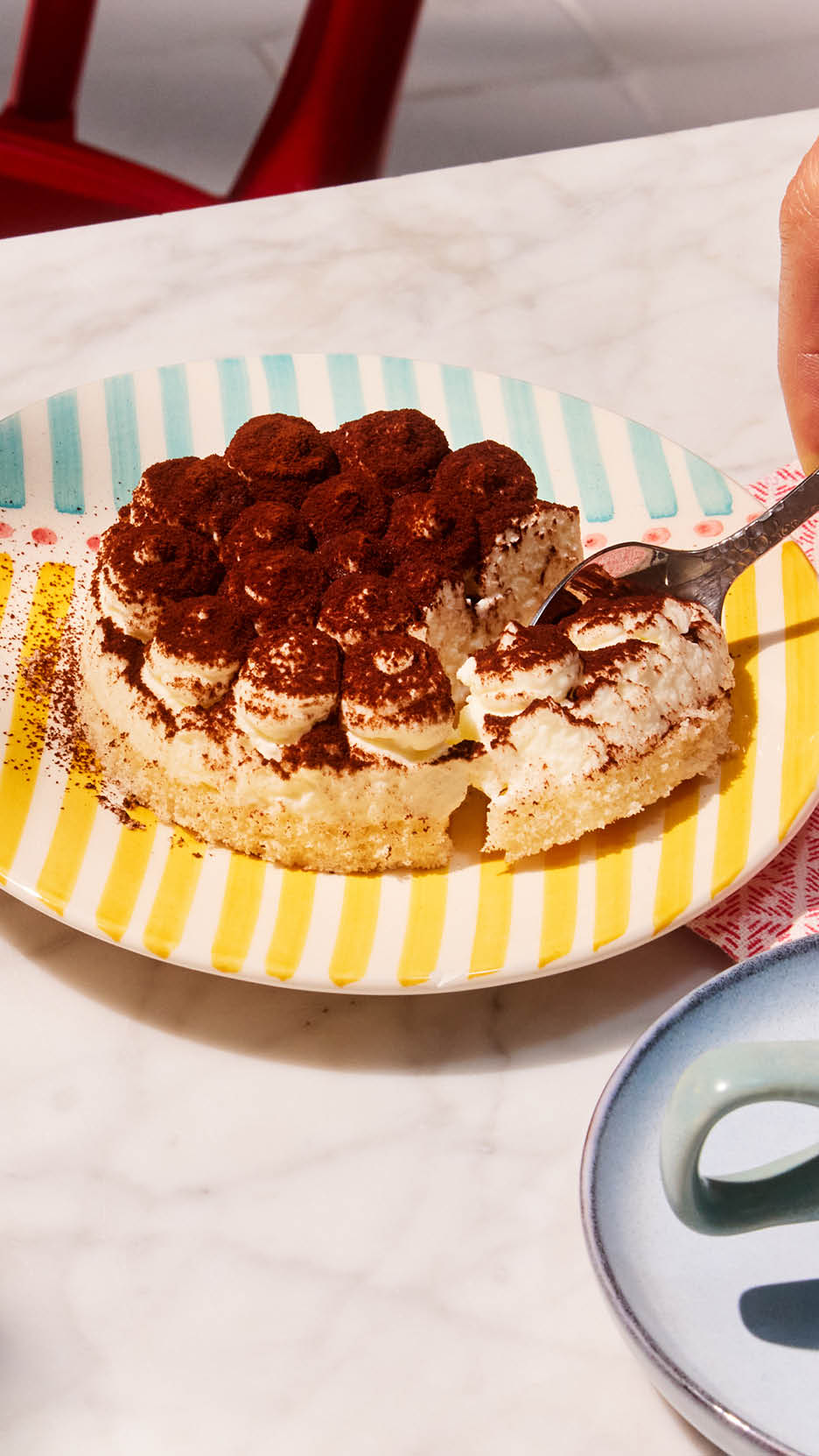 A tiramisu cake with cocoa powder on a striped plate, being served with a spoon.