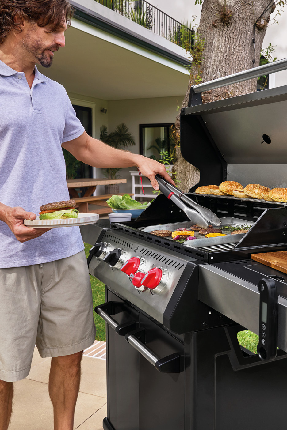Man grilling burgers on an outdoor gas grill, holding a plate with a finished burger.