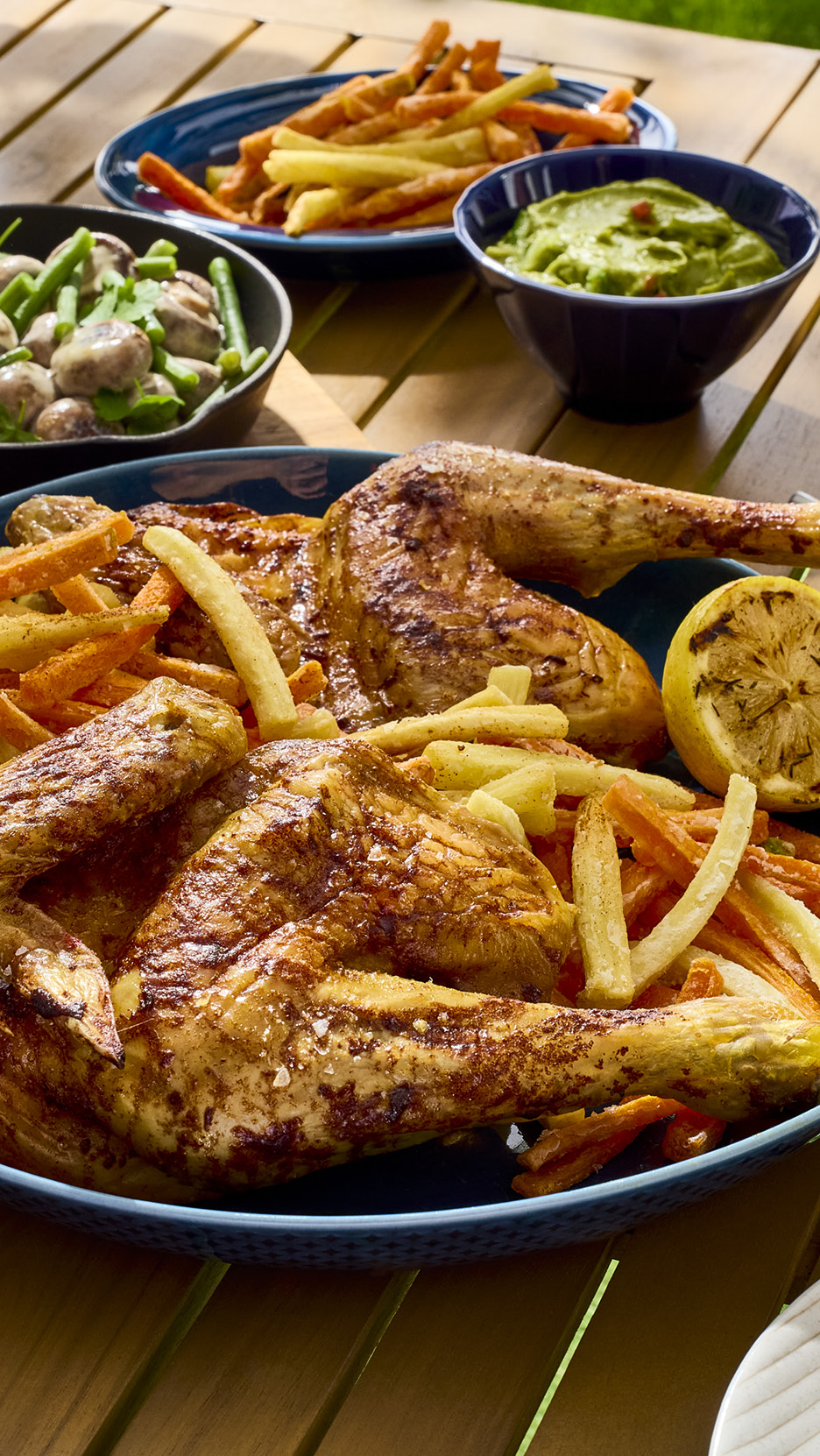 Grilled chicken with fries, mushrooms, green beans, and guacamole on a wooden table.