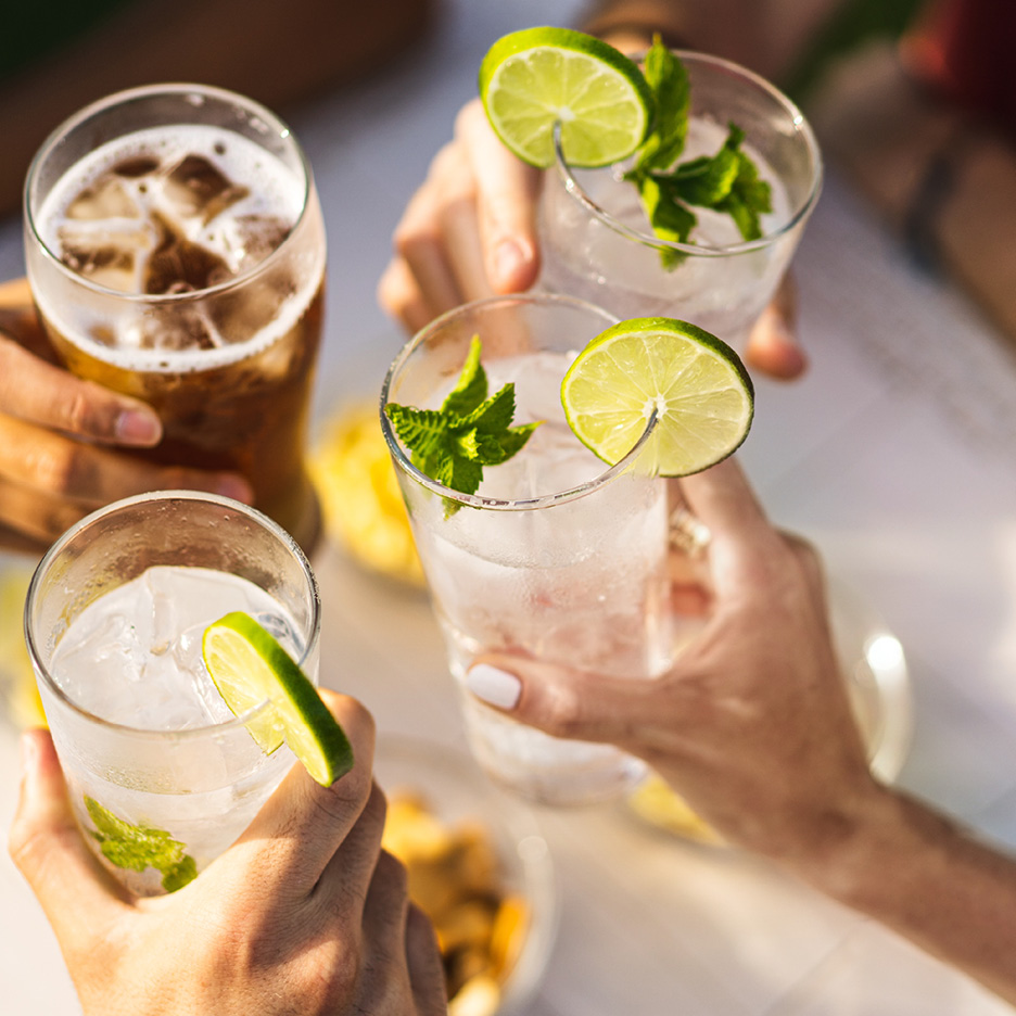 Hands holding glasses of beer and cocktails with lime and mint, enjoying a sunny day.