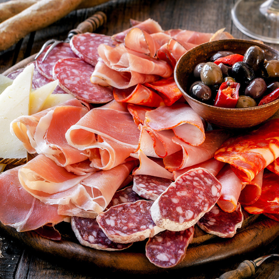 A charcuterie board with various cured meats, cheeses, and a bowl of olives and peppers.