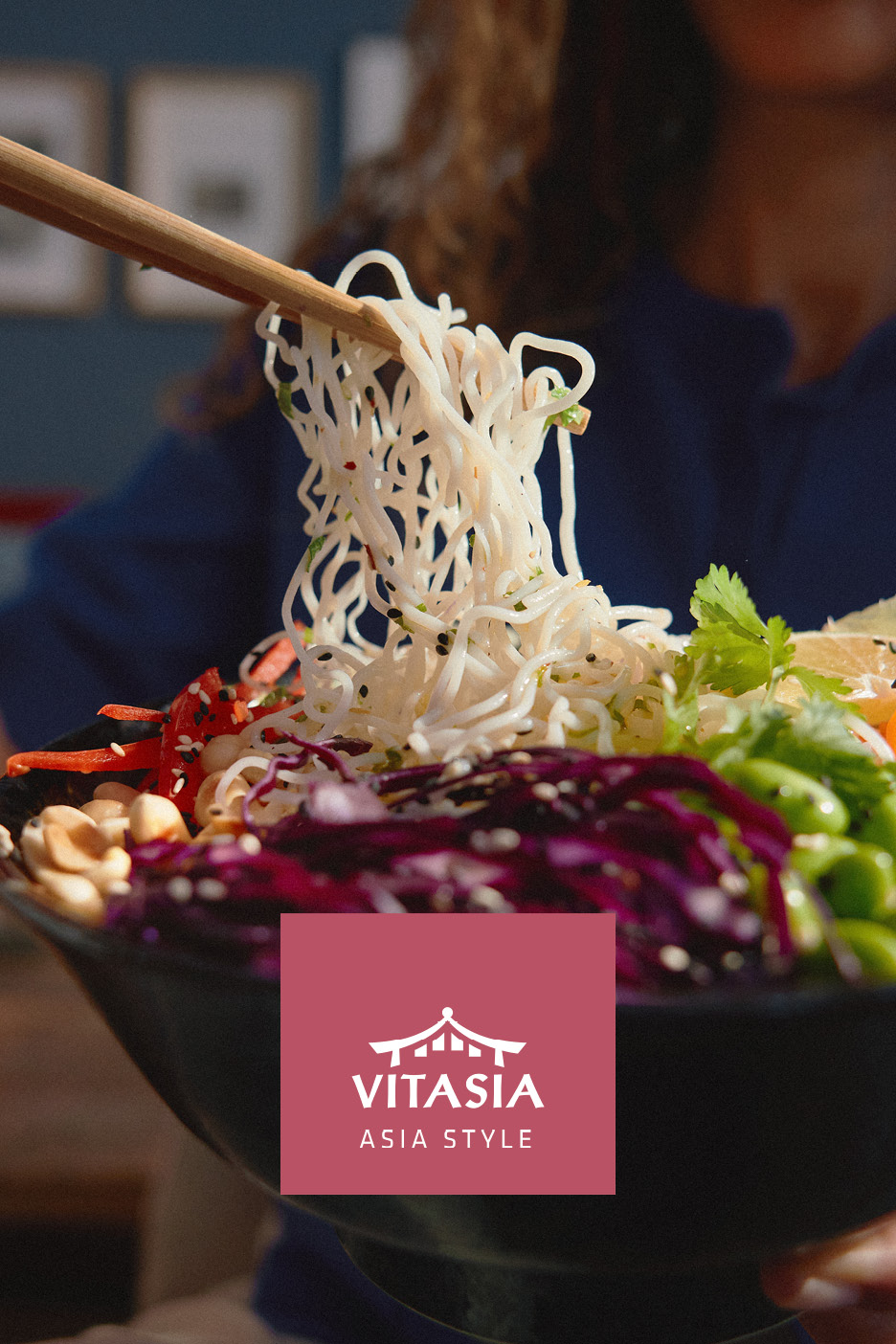 Chopsticks lifting noodles from a colorful Asian-style bowl with vegetables and peanuts.