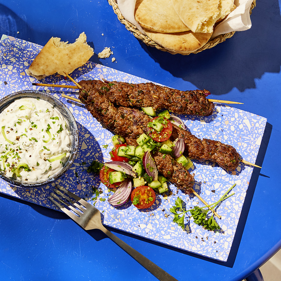 Kofta kebabs with tzatziki, tomato and cucumber salad, and flatbread on a blue table.