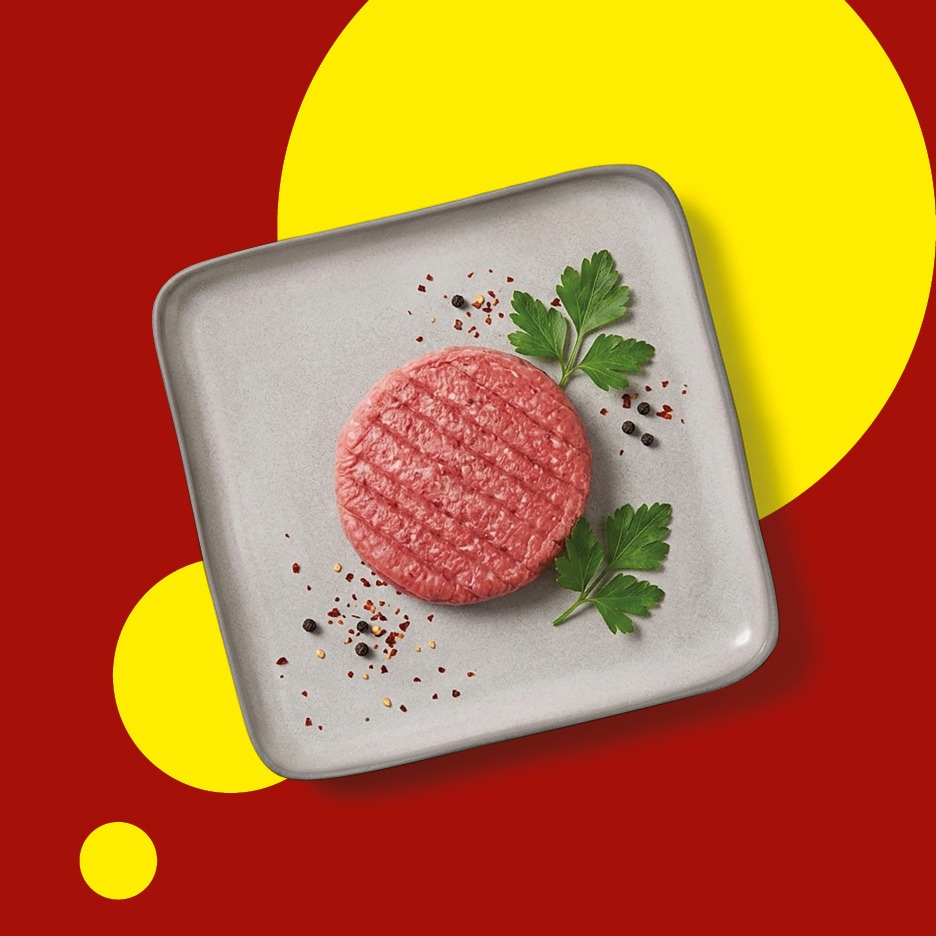 Raw burger patty on a grey plate with parsley and spices, on a red and yellow background.