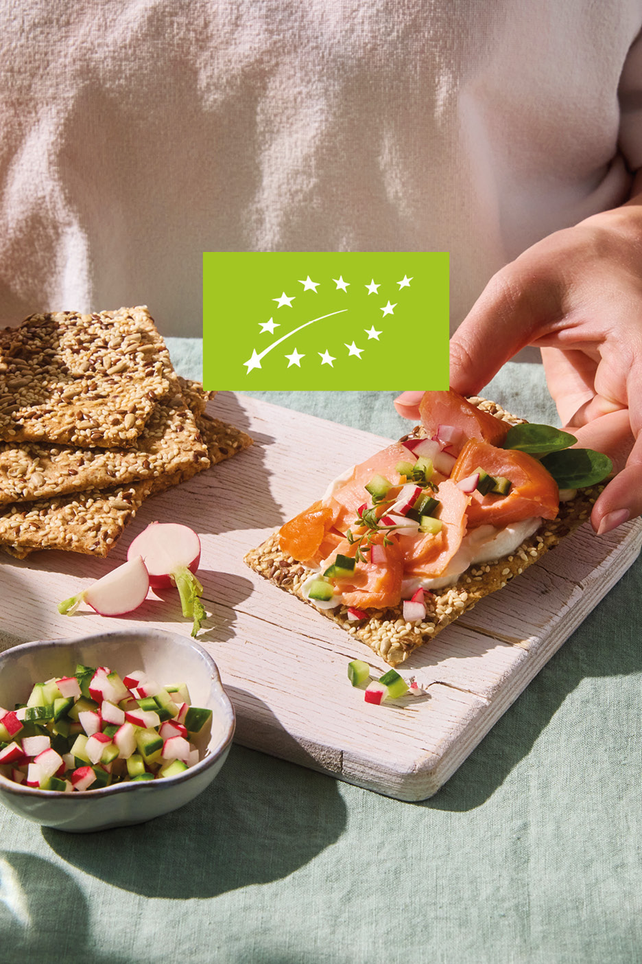 Hand placing smoked salmon and vegetable topping on a crispbread, with an organic label.