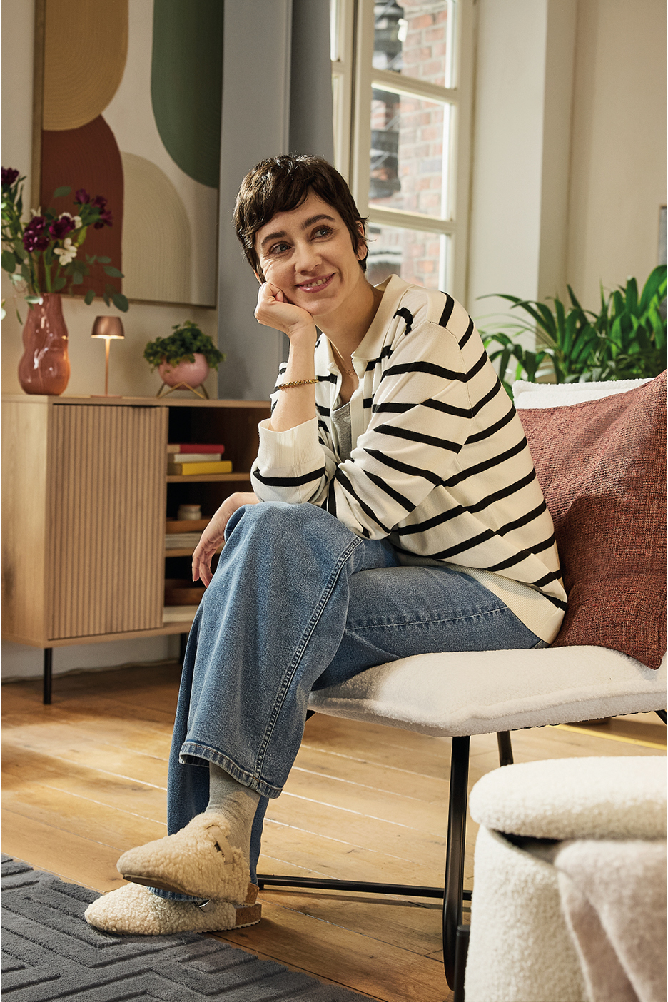 A woman in a striped sweater and jeans sits on a chair, wearing fluffy slippers.