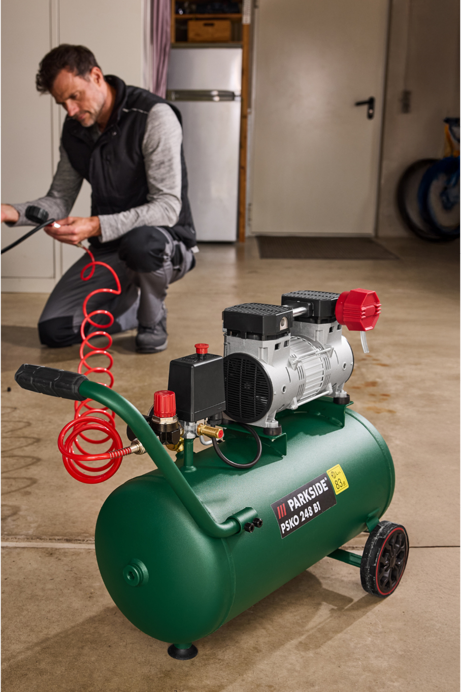 A man kneels in a garage, connecting a red coiled hose to a green Parkside air compressor.