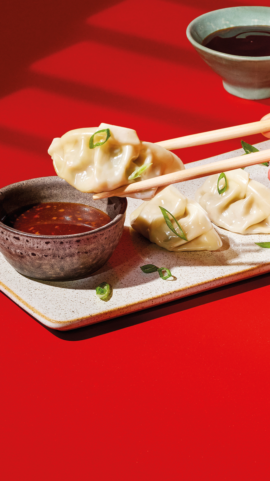 Chopsticks holding a dumpling over a bowl of chili oil, with more dumplings on a plate.