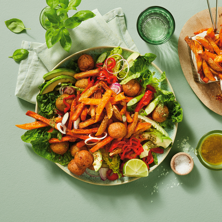 A vibrant salad with sweet potato fries, falafel, avocado, and red peppers on a green background.