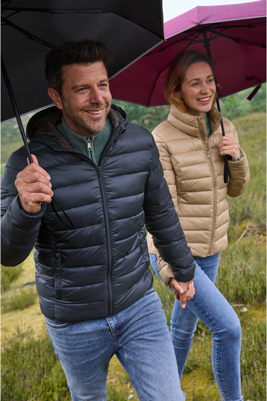 A couple in puffer jackets and jeans holding umbrellas and hands, walking outdoors.