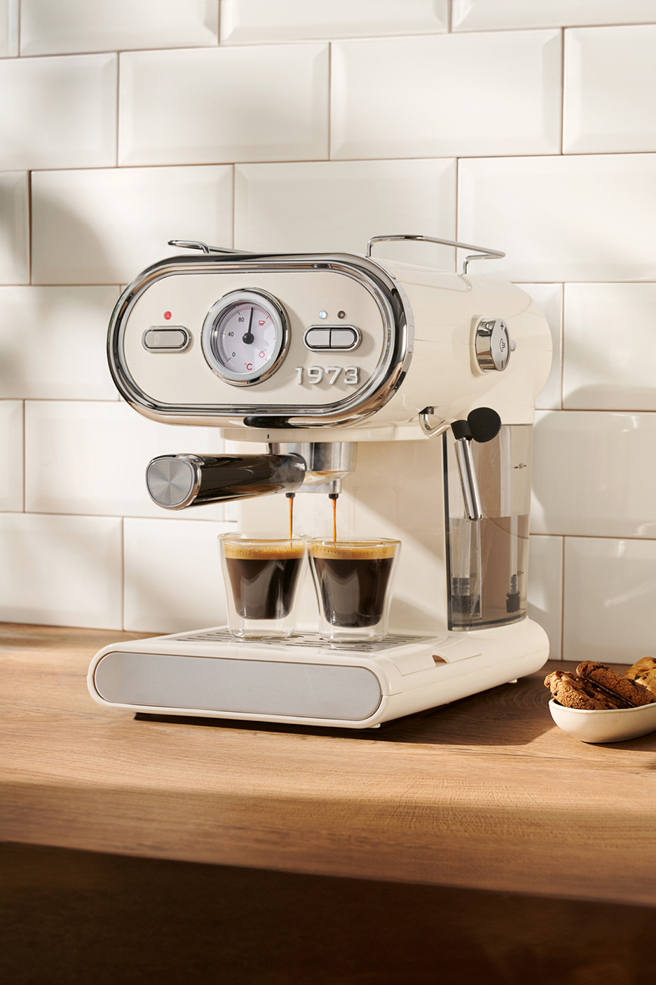 Cream-colored espresso machine brewing two shots of coffee into clear glasses on a wooden counter.