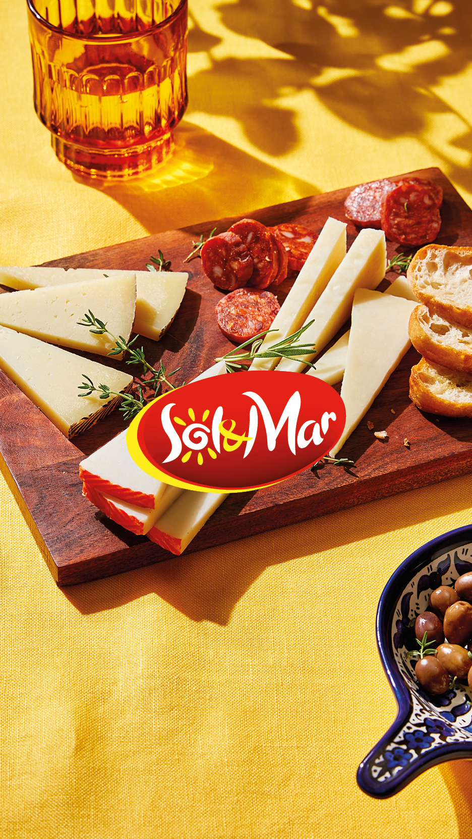 A wooden board with Sol & Mar cheese, chorizo, and bread, next to a glass and olives.