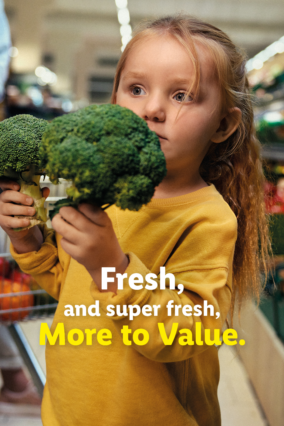 A young girl in a supermarket holding two heads of broccoli.