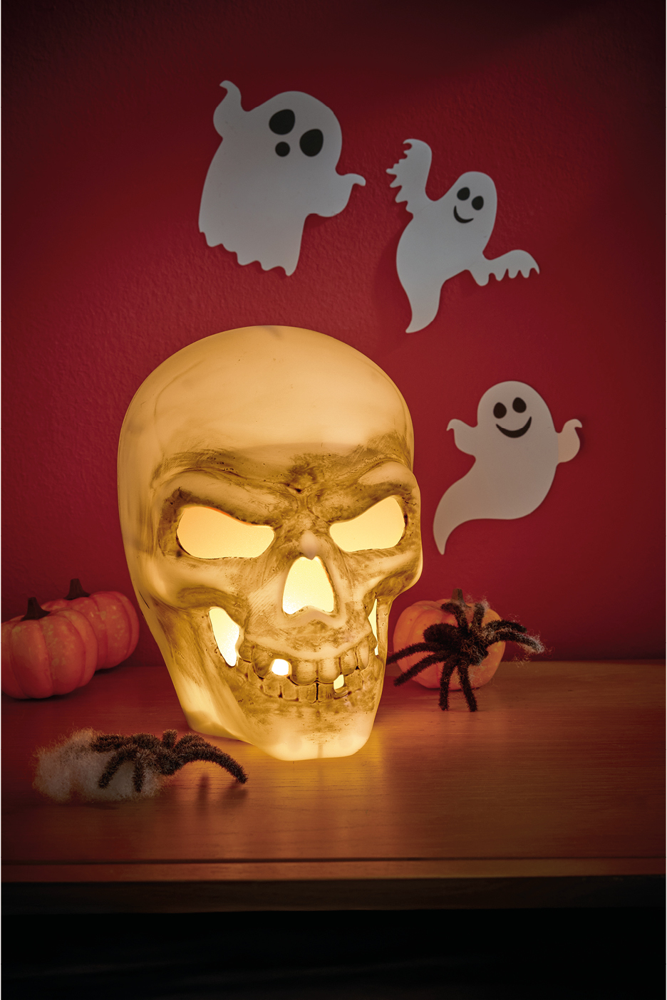 A glowing skull light, pumpkins, and spiders on a table with ghost cutouts on a red wall.