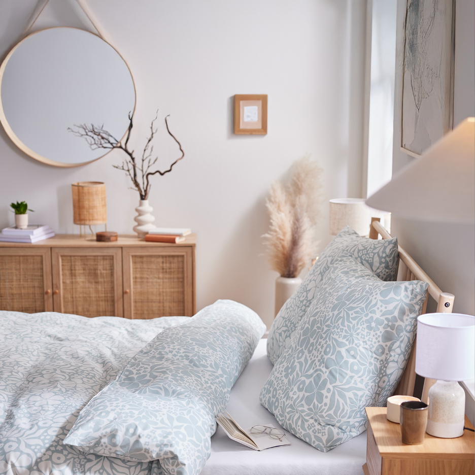 Bedroom with light blue floral duvet cover and pillows, wooden furniture, and a round mirror.