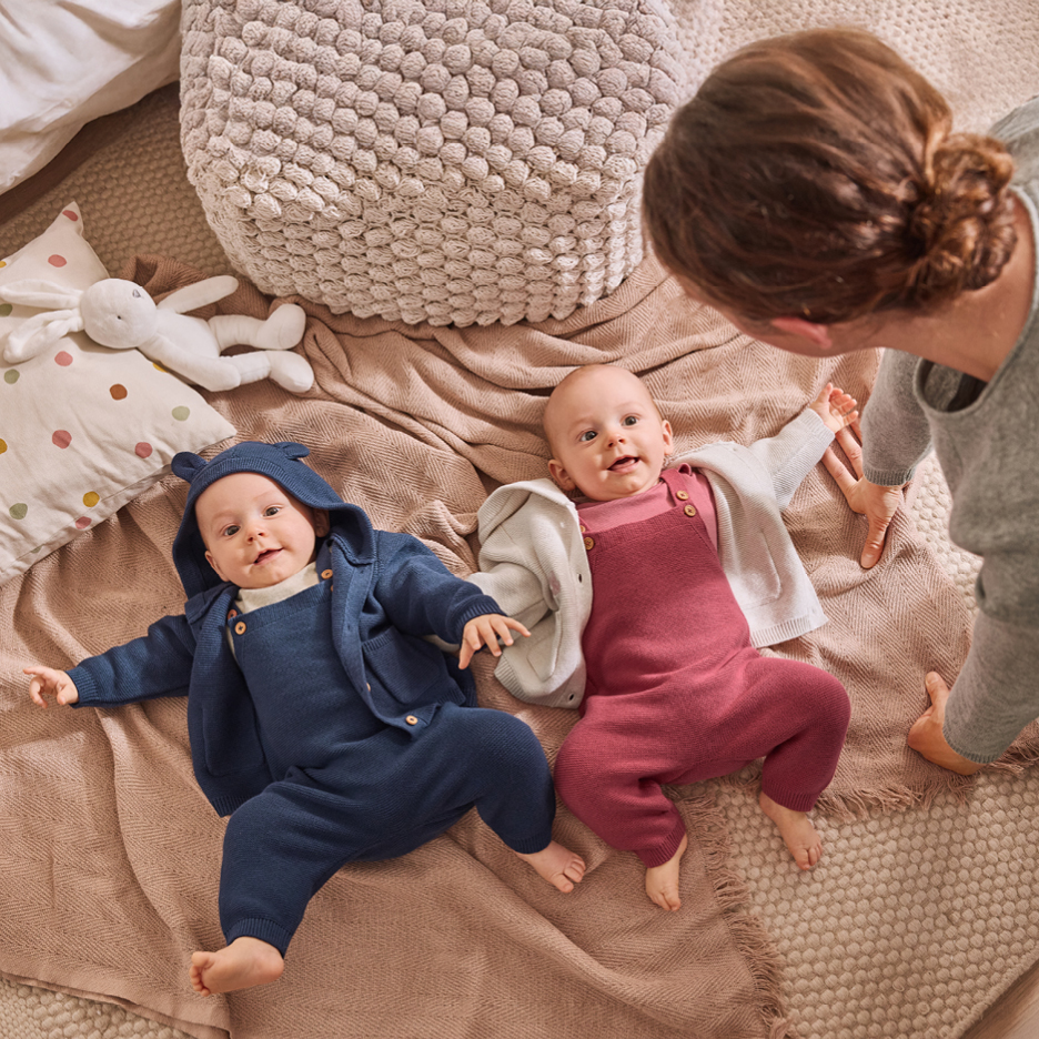 Two babies in knit rompers and cardigans on a blanket, with an adult nearby.