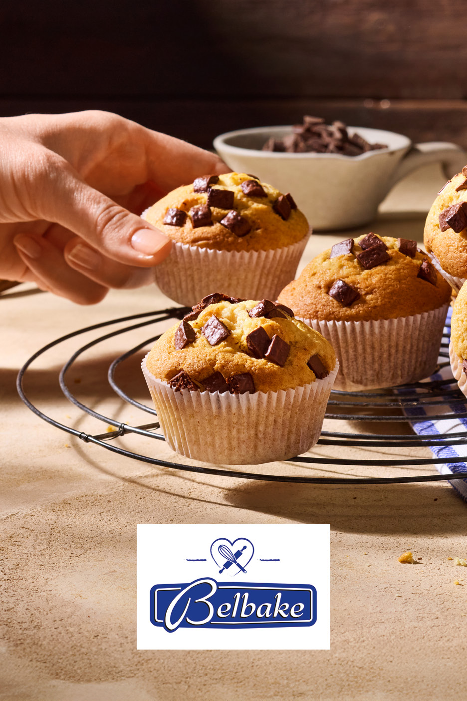 Freshly baked chocolate chip muffins on a cooling rack with a hand reaching for one.
