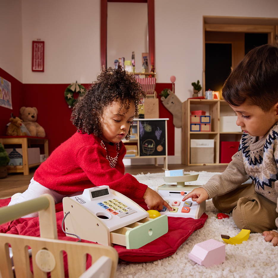 Two children playing with a wooden toy cash register and scale on a rug.