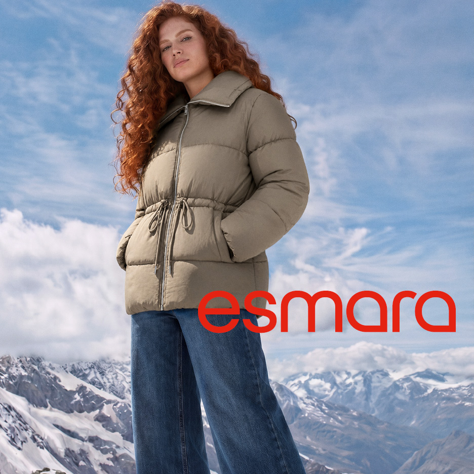 A woman in a beige puffer jacket and blue jeans stands against a snowy mountain backdrop.