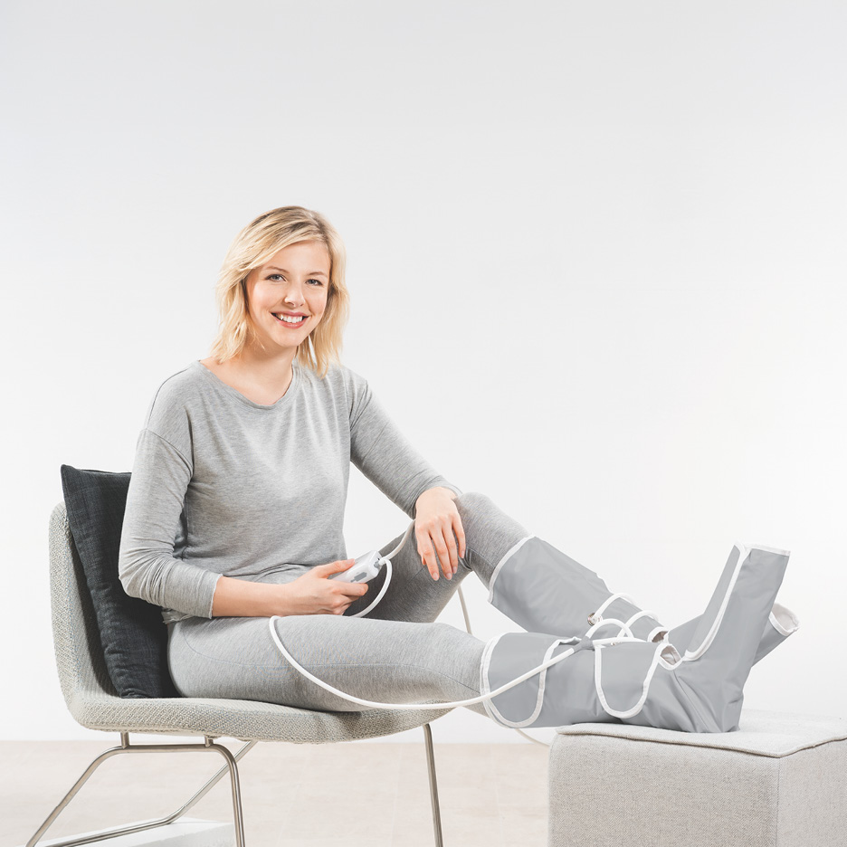 A woman using a grey leg compression massager while sitting on a chair.