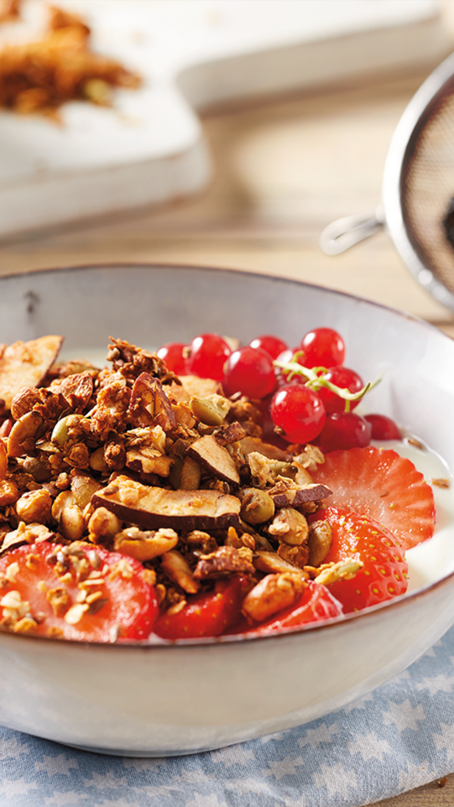 Granola with strawberries, red currants, and yogurt in a white bowl on a patterned cloth.