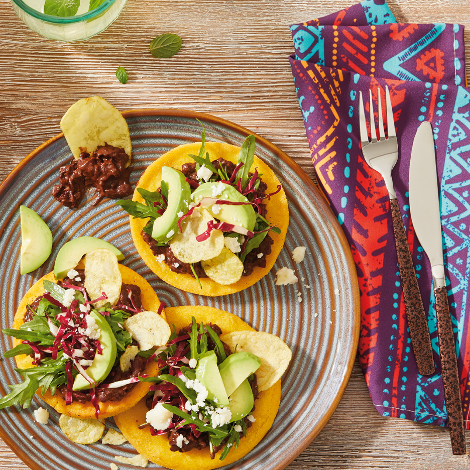 Three corn tortillas with black beans, avocado, arugula, and potato chips on a plate.