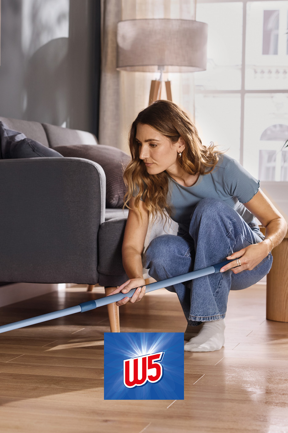 A woman cleaning under a sofa with a flat mop on a wooden floor.