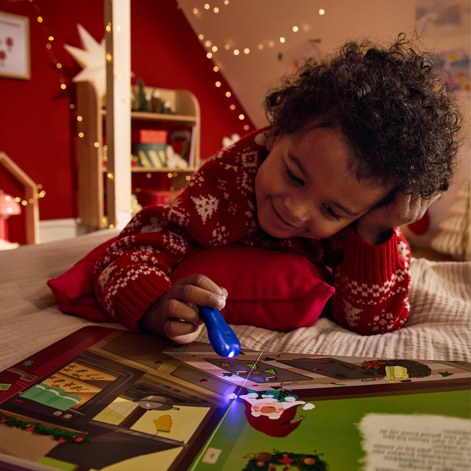 Smiling child in a Christmas jumper reading a book with a light pen on a bed.