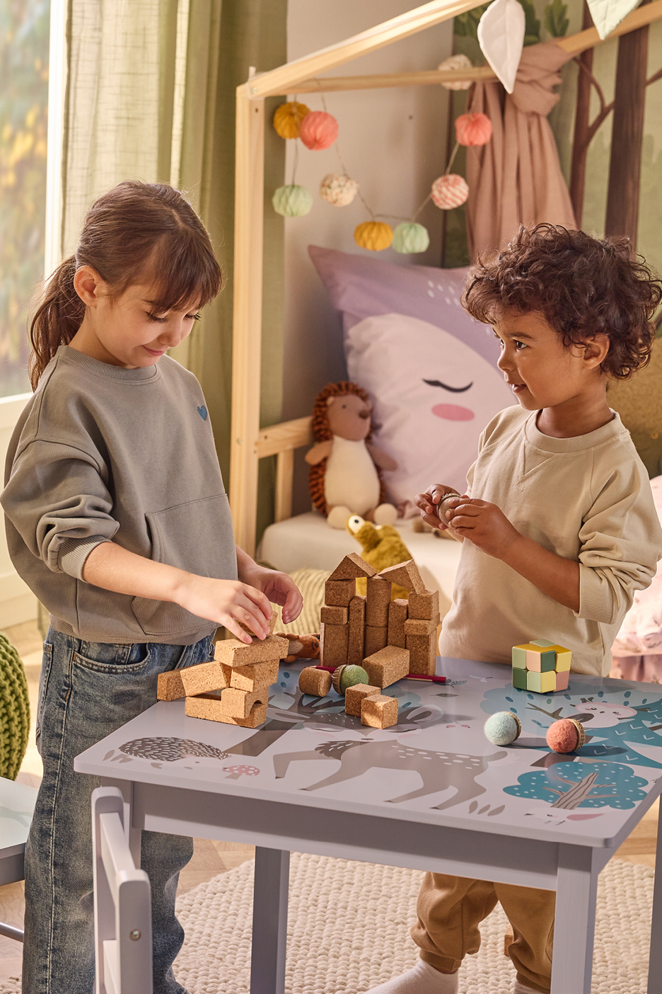 Two children playing with cork building blocks and other toys on a patterned table.