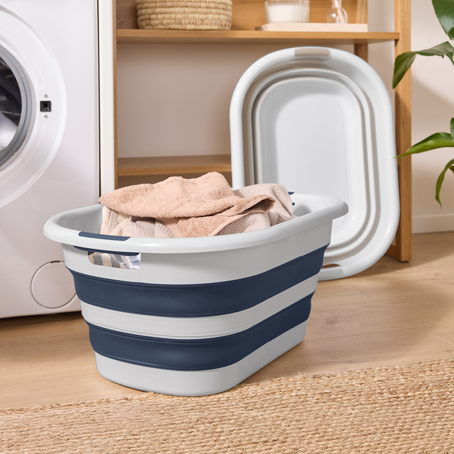 Foldable laundry basket with clothes next to a washing machine and a wooden shelf.