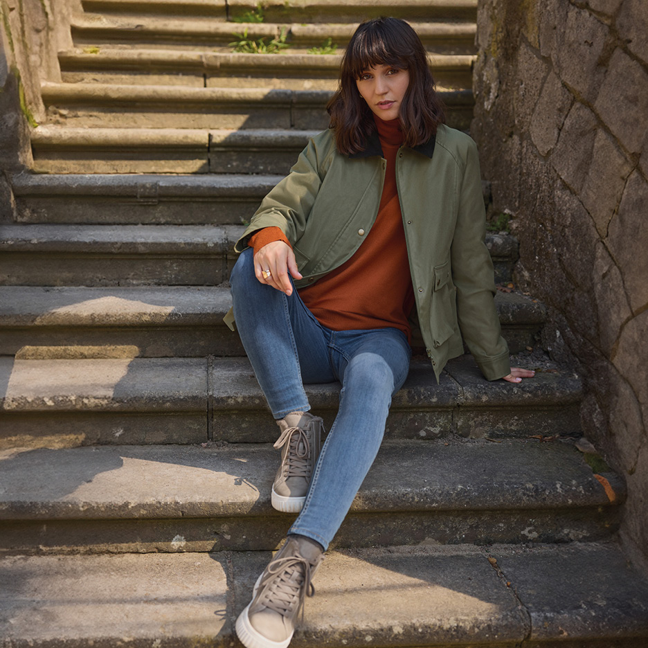 Woman in a green jacket, rust-colored turtleneck, and blue jeans sitting on stone steps.