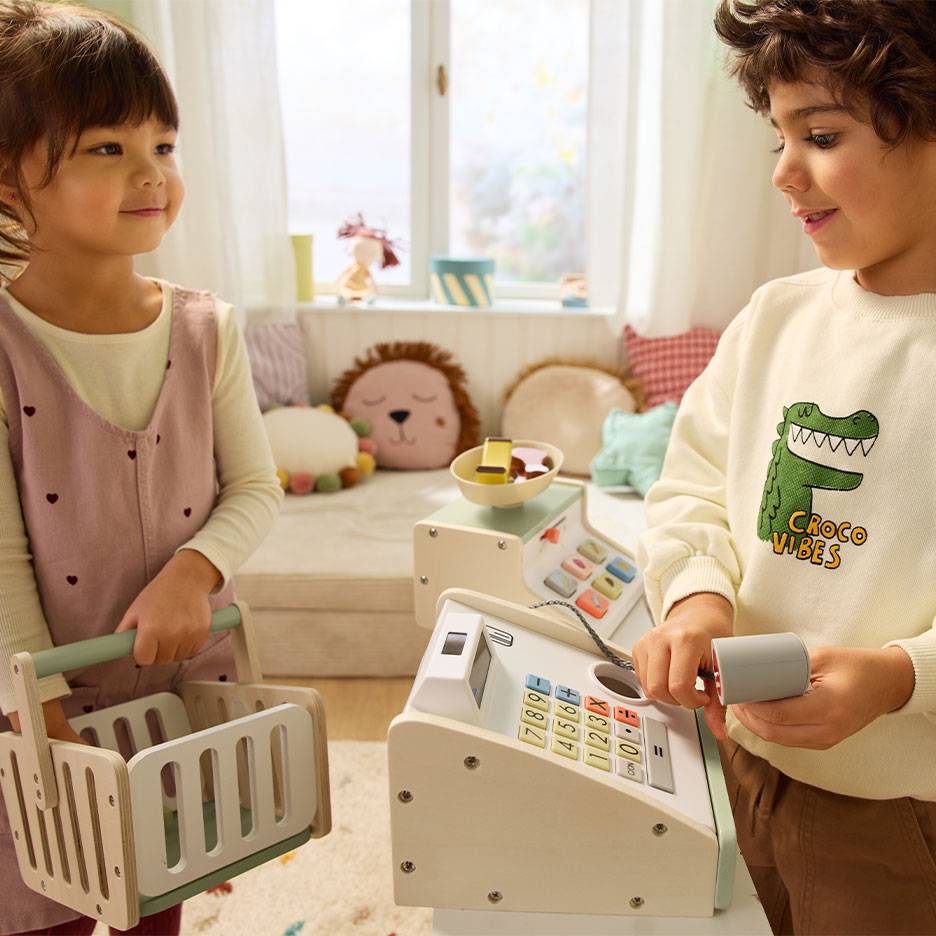 Children playing shop; boy operating a cash register, girl carrying a shopping basket.
