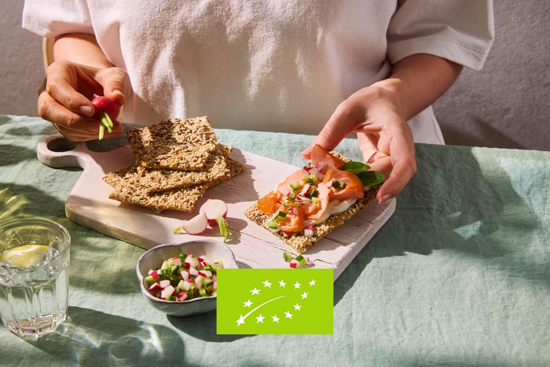 Hands preparing crispbread with salmon, cream cheese, and radishes on a light green tablecloth.