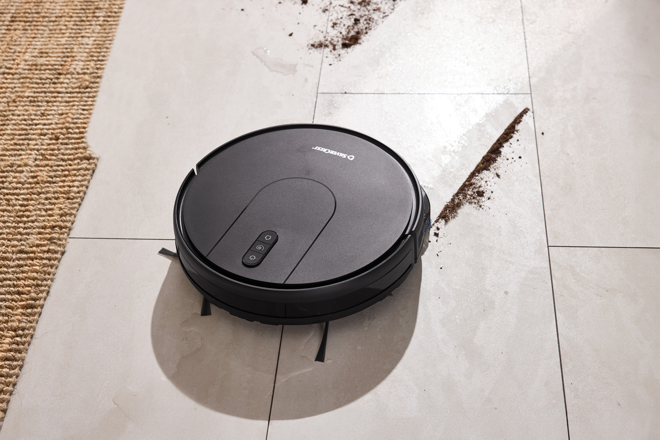 Silvercrest robot vacuum cleaner on a tiled floor with dirt and water, next to a rug.