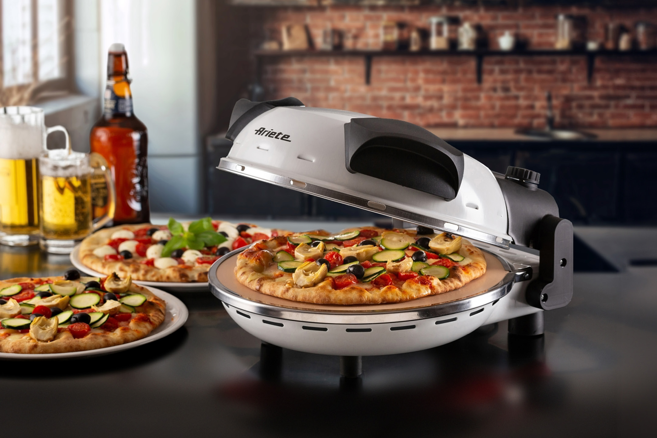 Ariete pizza oven with three pizzas, two beers, and a kitchen background.