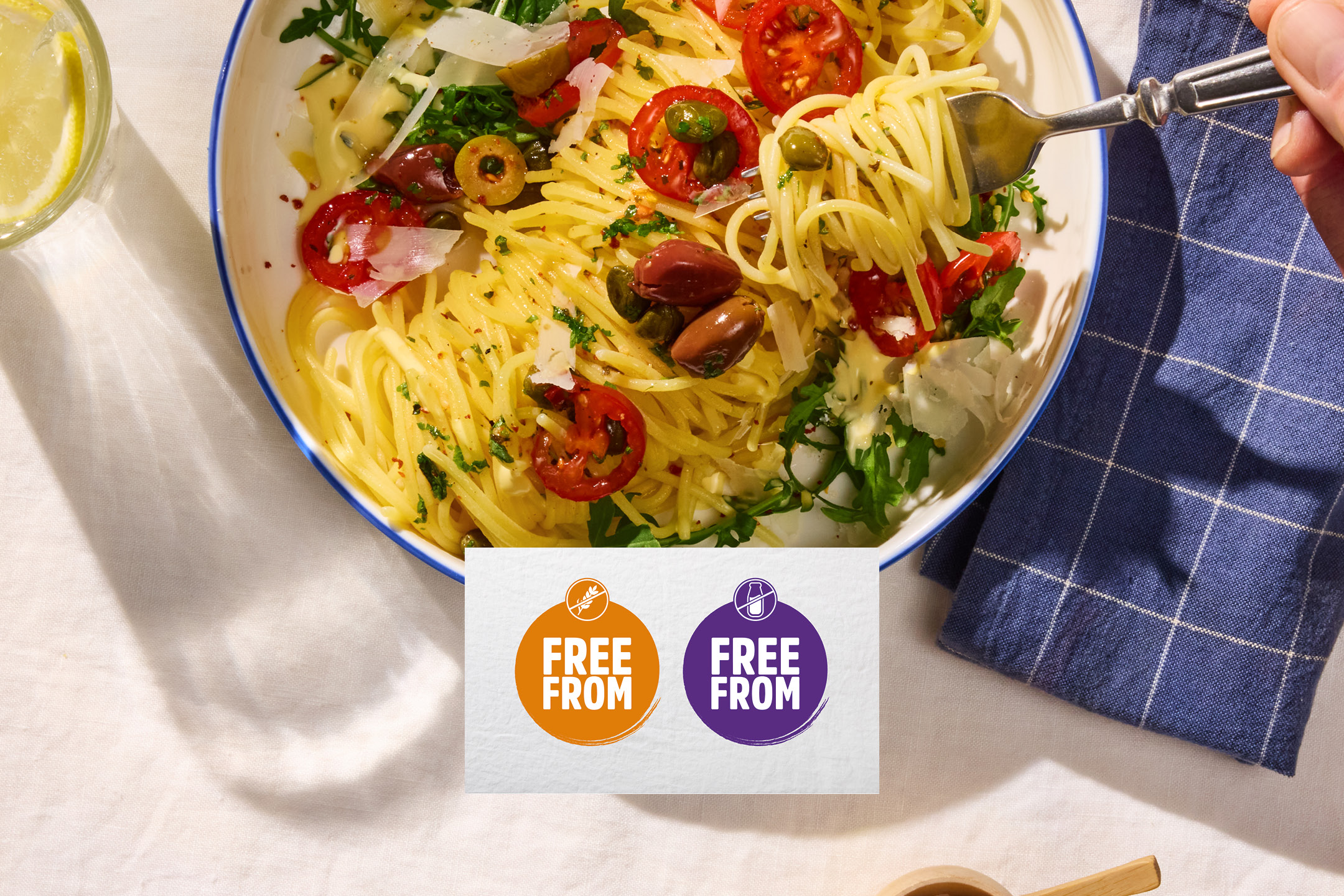 Overhead shot of spaghetti with cherry tomatoes, olives, capers, and parmesan, with 'FREE FROM' labels.