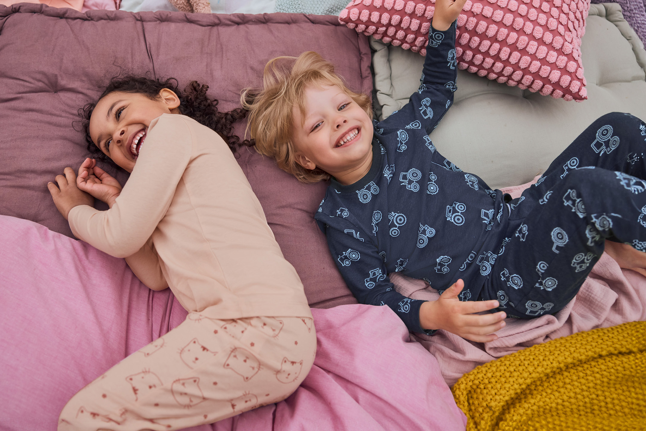 Two happy children in pajamas lying on colorful bedding and pillows.