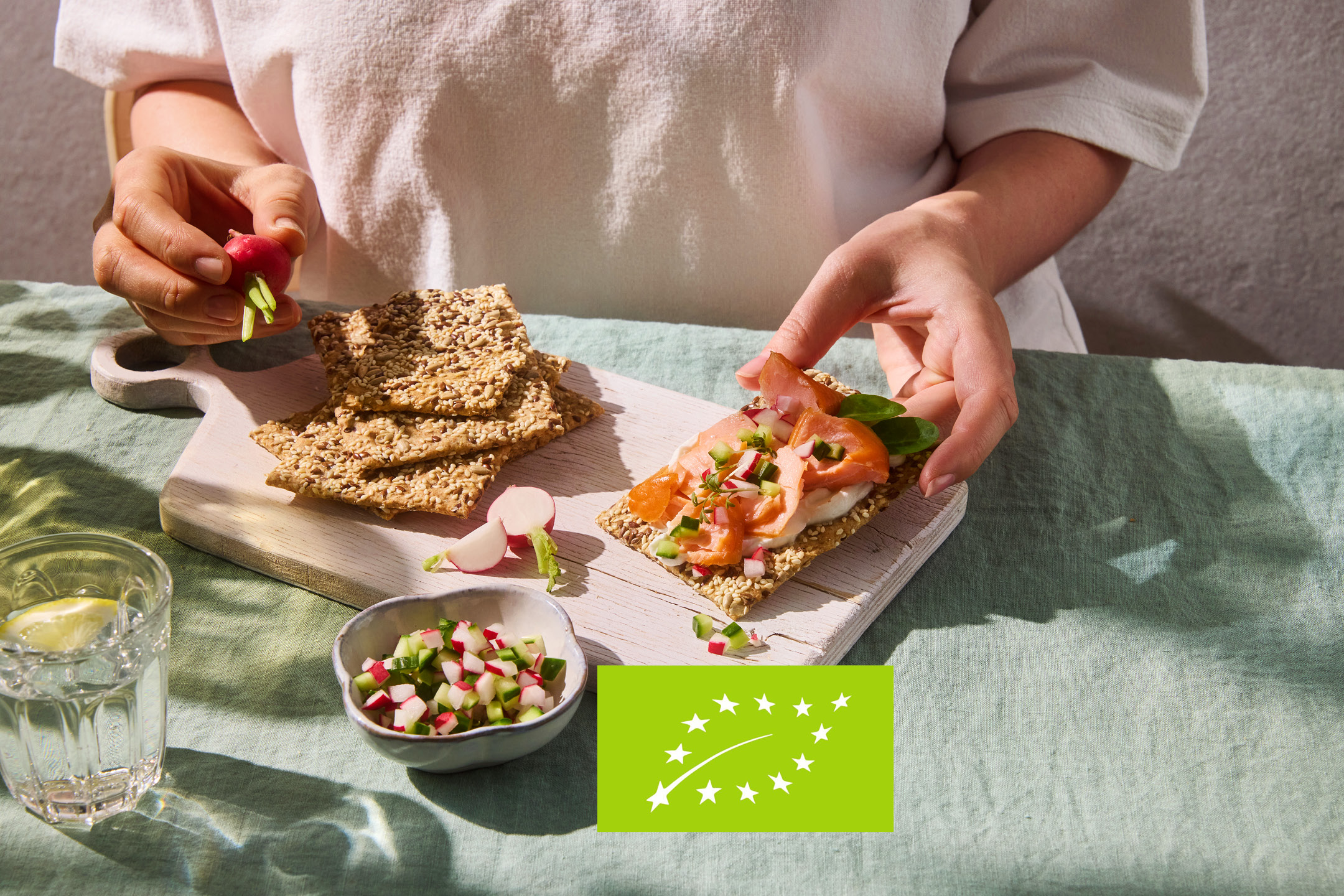 Hands preparing a crispbread with salmon, cream cheese, and vegetables on a wooden board.