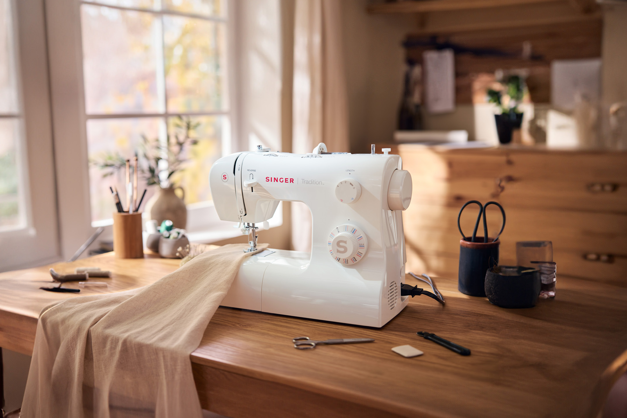 White SINGER Tradition sewing machine on a wooden table with fabric and sewing accessories.