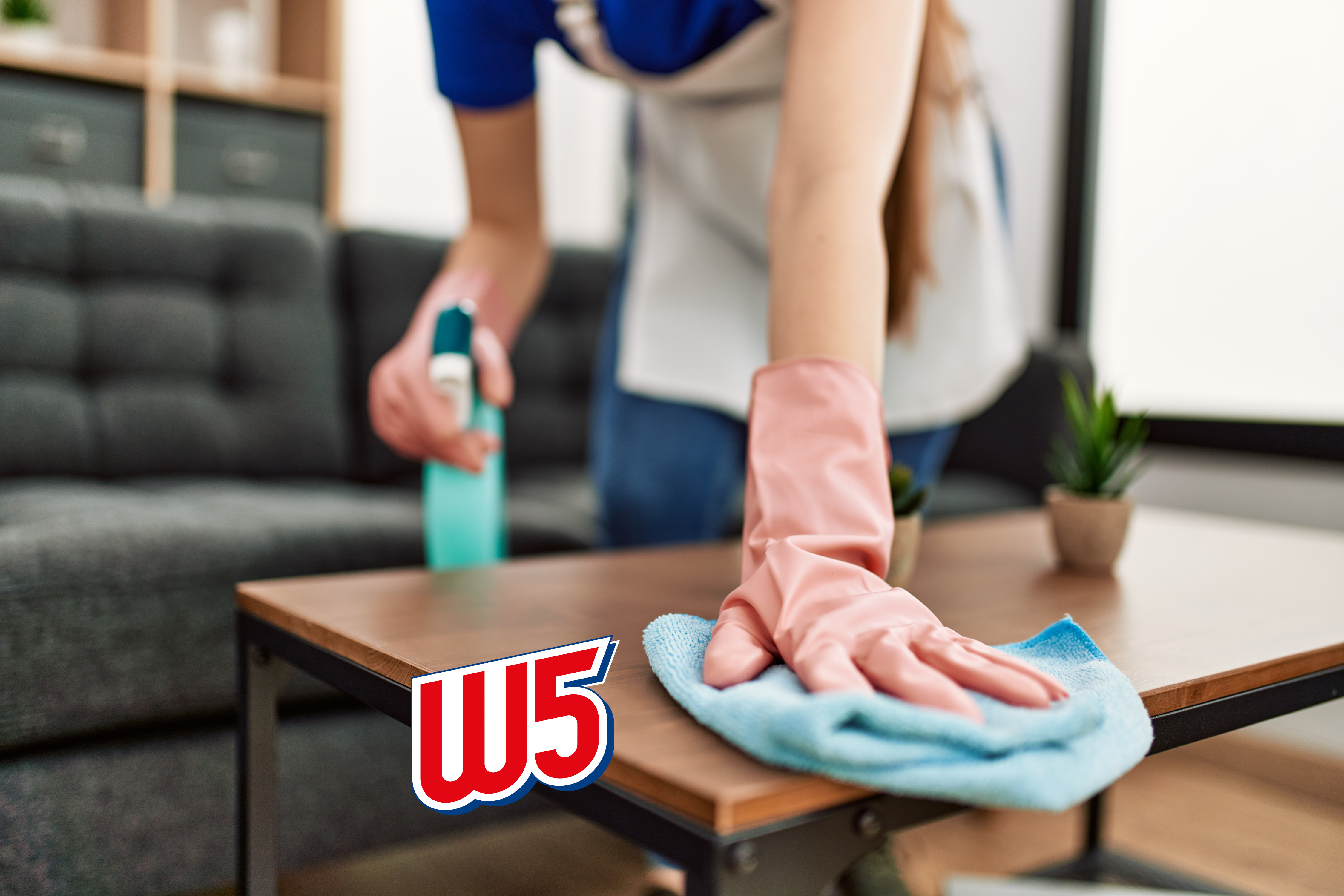 Person cleaning a wooden table with a spray bottle and a cloth, wearing pink gloves.