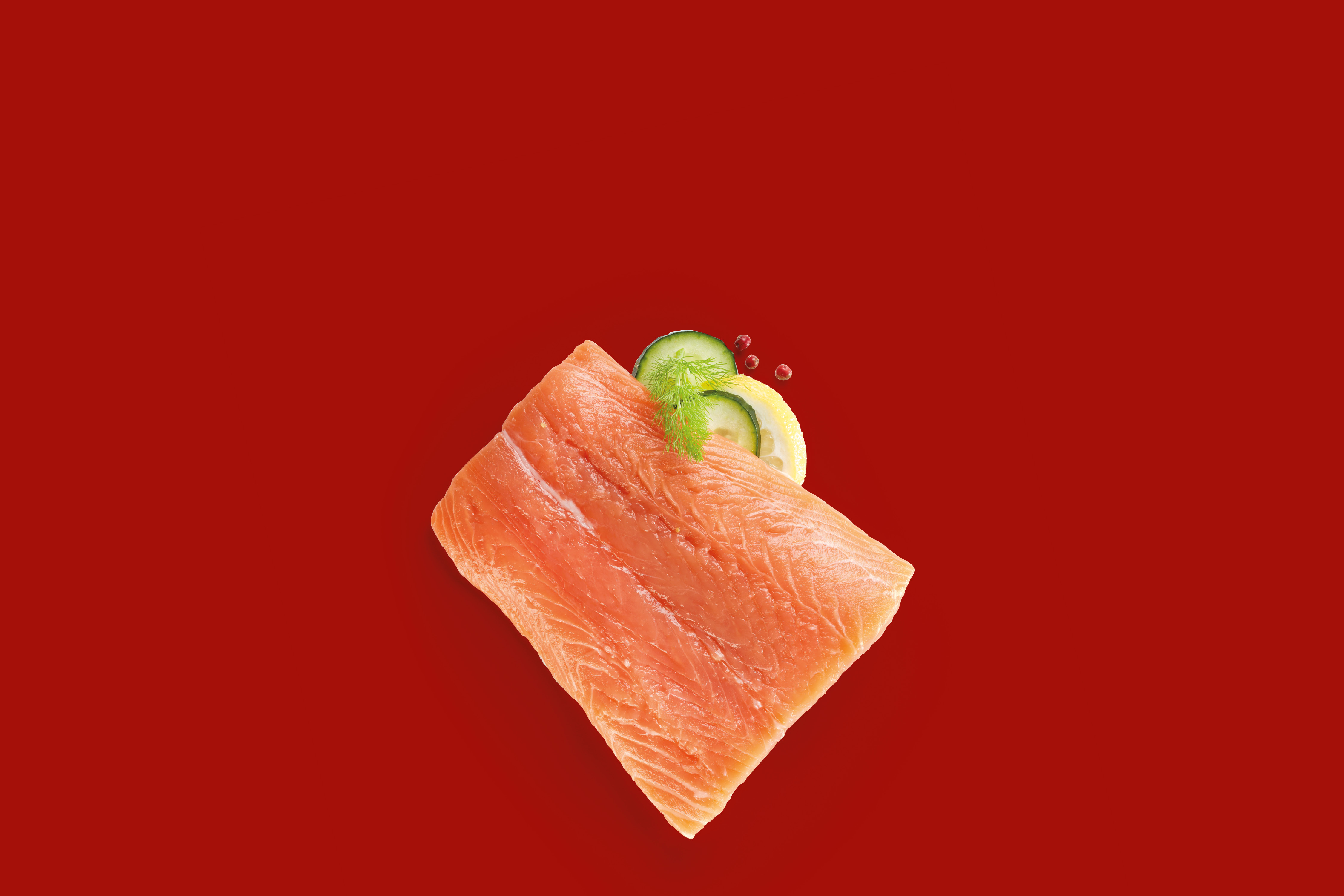 Fresh salmon fillet with cucumber, lemon, dill, and pink peppercorns on a red background.