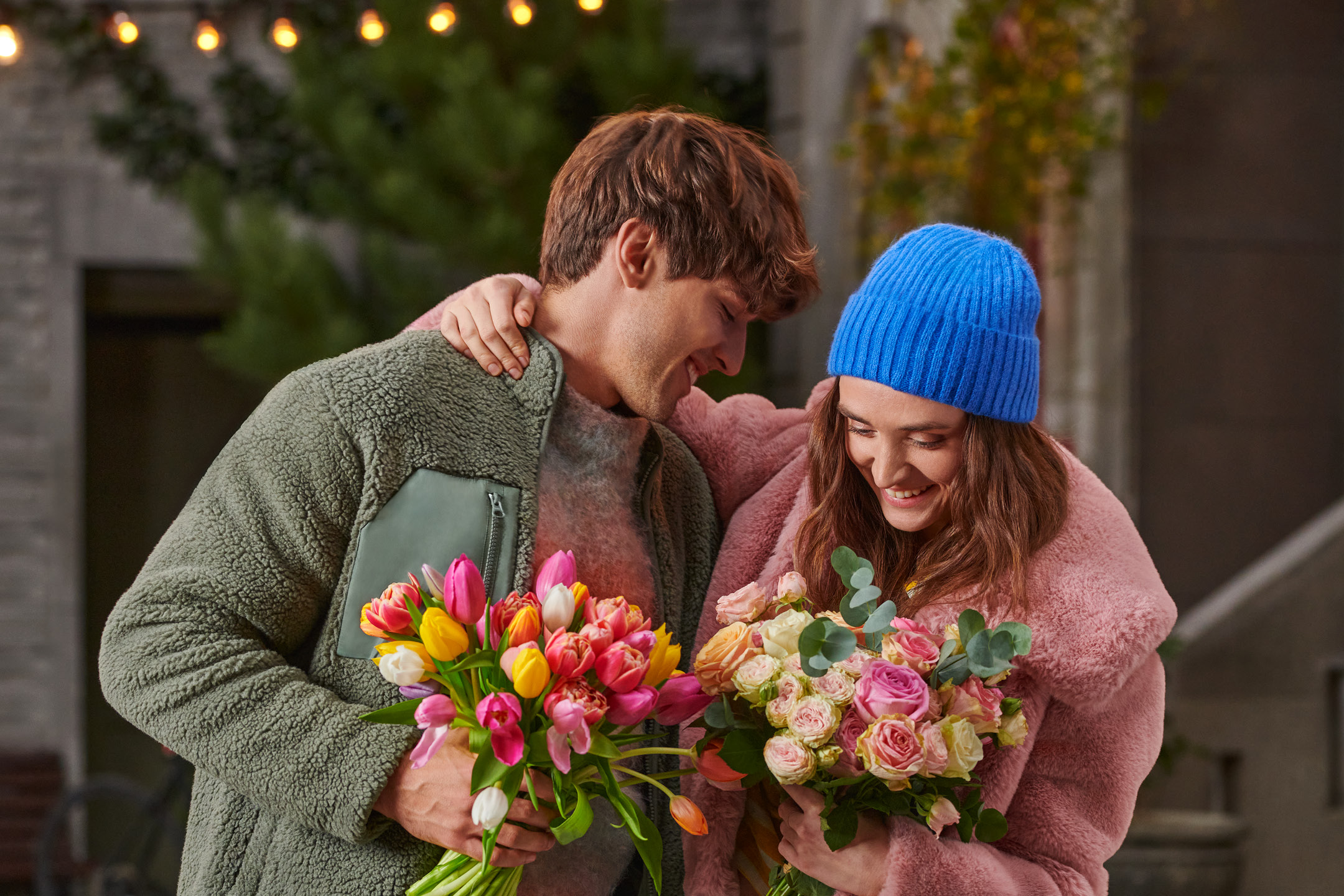 Couple in cozy jackets holding bouquets of colorful tulips and roses, smiling warmly.