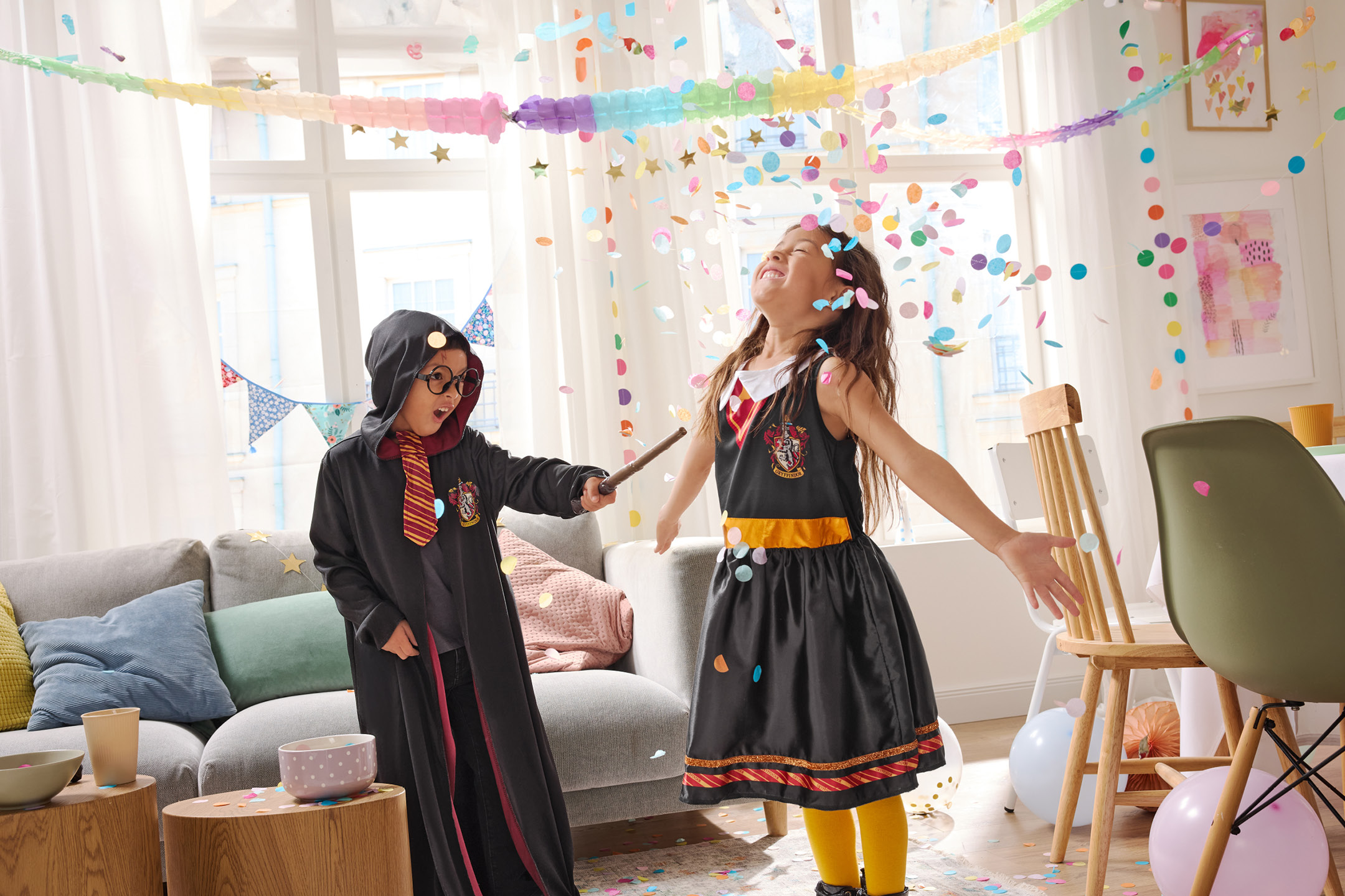 Two children in wizard costumes playing with confetti in a decorated living room.